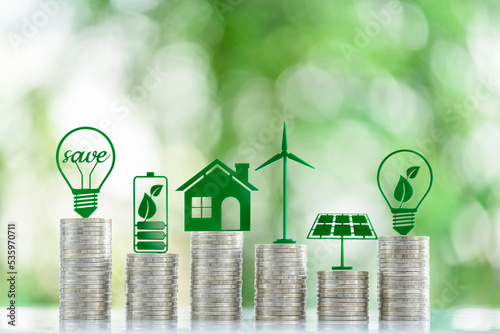 Renewable or clean energy generation prices and costs, financial concept : Green eco-friendly symbols atop coin stacks e.g. energy efficient light bulb, a battery, a solar cell panel, a wind turbine.