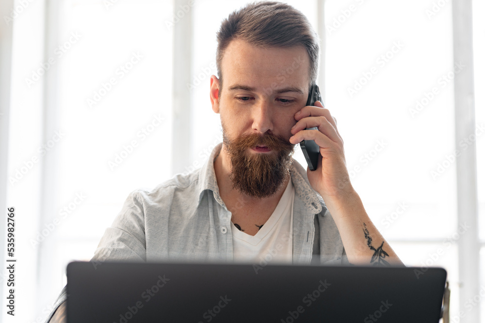 Young bearded man working from home with laptop while talking on the phone Young bearded man working from home with laptop while talking on the phone