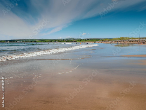 Yellow sand and blue ocean and sky. People swim in the background and surf on board. Lahinch beach in county Clare, Ireland. Warm sunny day. Irish landscape.