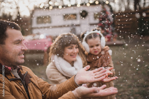Happy family with two daughters dressed in stylish fur coats a play hugging and enjoying near the trailer in the forest with New Year's decorations. Selected focus