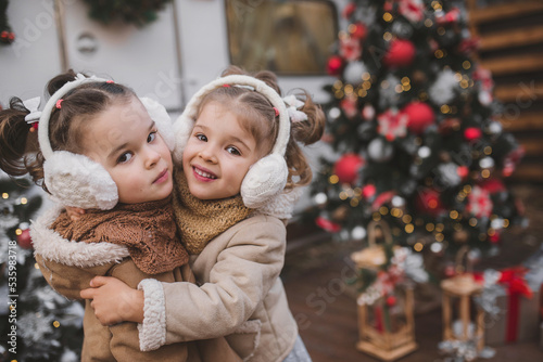 two little twin girls dressed in stylish fur coats play  hug and enjoy near the trailer in the forest with New Year s decorations. Family decorating the Christmas tree
