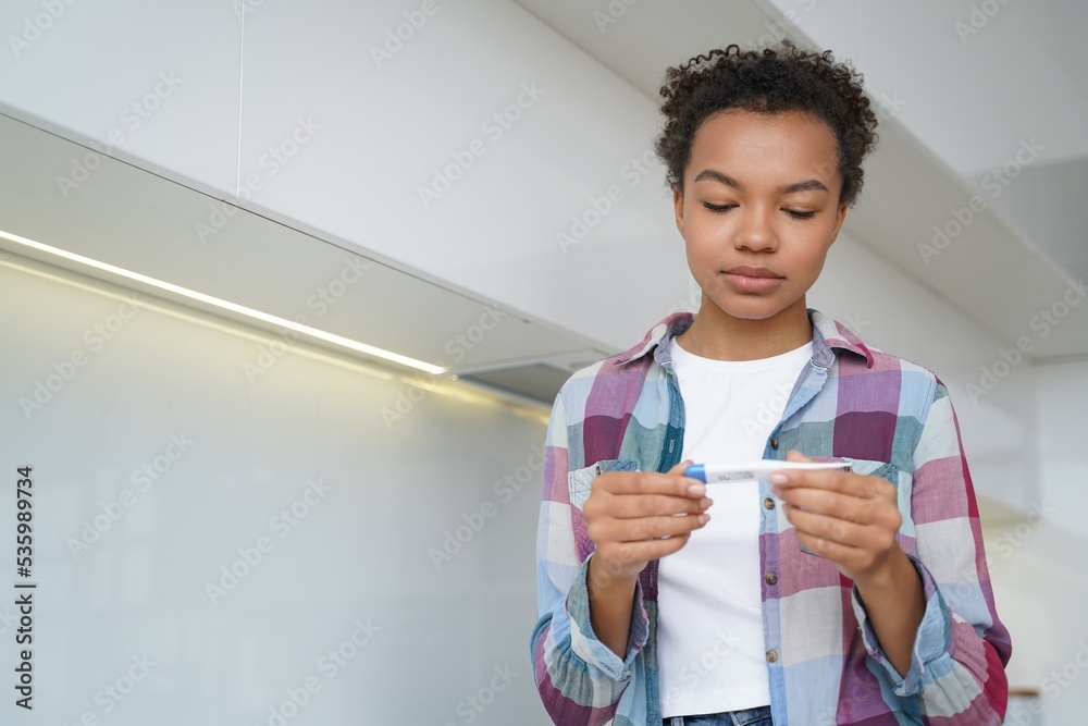 Unhealthy mixed race teen girl check body temperature with digital thermometer, standing at home Unhealthy mixed race teen girl check body temperature with digital thermometer, standing at home
