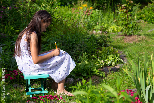 Young caucasian girl in a white dress is drawing on a plein air sitting on a folding chair in summer garden 