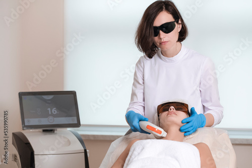 Professional Cosmetologist Treating Human Skin with Laser While Touching of Female Face with Apparatus for Intense Pulse Light or IPL photo