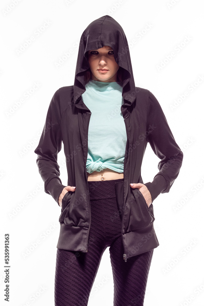 Natural Portrait of Winsome Sportive Caucasian Female Athlete Posing In Black Hoody and Sexy Fitness Outfit While Looking Straight Against White Background. Natural Portrait of Winsome Sportive Caucasian Female Athlete Posing In Black Hoody and Sexy Fitness Outfit While Looking Straight Against White Background.