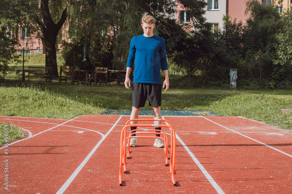Blond boy in sportswear jumps over red obstacles to improve lower body dynamics. Plyometric training in an outdoor environment. Improve your skills