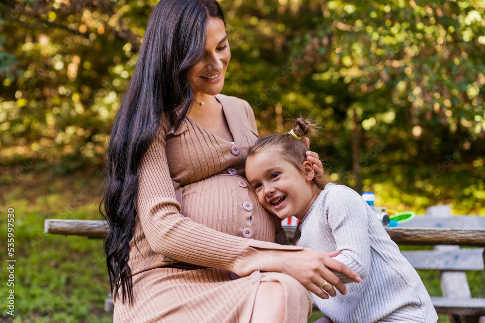 Happy pregnant mother and daughter hugging on a park bench. Happy motherhood. Beautiful woman with a little daughter in nature Happy pregnant mother and daughter hugging on a park bench. Happy motherhood. Beautiful woman with a little daughter in nature