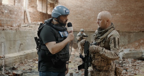 POV War journalist correspondent wearing bulletproof vest and helmet reporting live near destroyed building, conducting interview with military representative