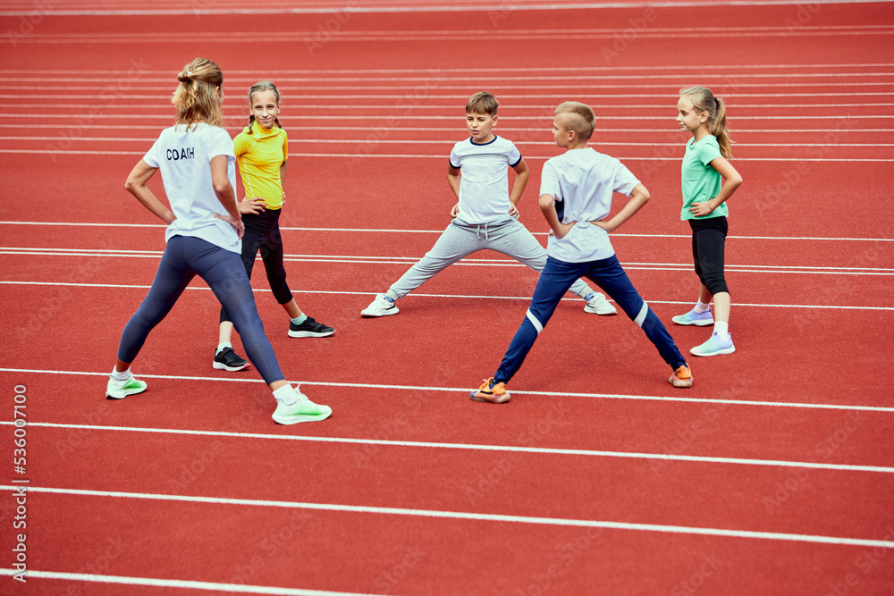 Fototapeta premium Female coach training athletes. Group of children running on treadmill at the stadium. Concept of sport, achievements, studying, goals, skills. Little boys and girls training outdoor.