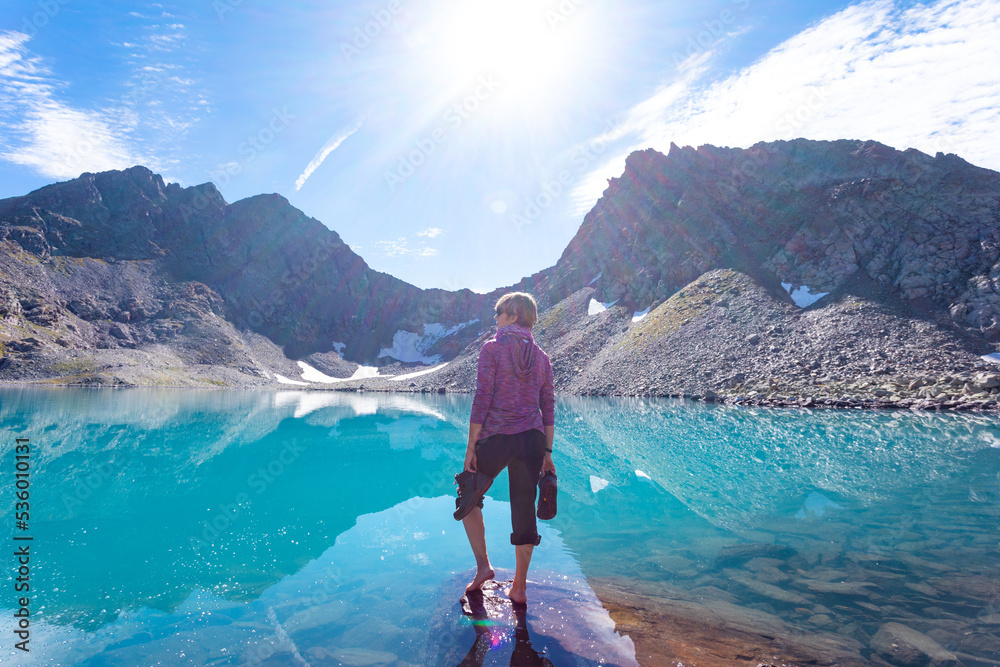 The girl is resting with bare feet against the backdrop of mountain peaks and a blue turquoise lake. He is holding shoes in his hands. Beautiful mountain landscape for recreation, travel and a healthy