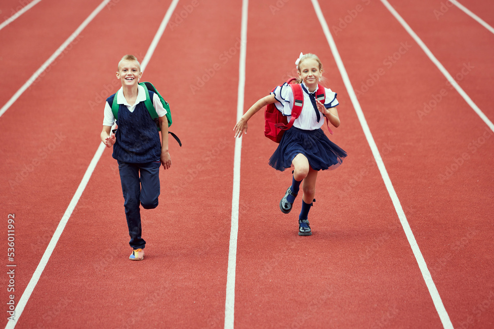 Start. Back to school, kids and education concept. Girl and boy dressed in school uniform as elementary student carrying big backpack running on treadmill at the stadium or arena.