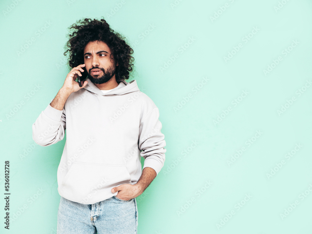 Handsome smiling hipster model.Unshaven Arabian man holding smartphone. Fashion male with long curly hairstyle posing near green wall in studio. Talking at call Handsome smiling hipster model.Unshaven Arabian man holding smartphone. Fashion male with long curly hairstyle posing near green wall in studio. Talking at call