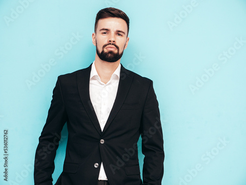 Portrait of handsome confident stylish hipster lambersexual model. Sexy modern man dressed in elegant black suit. Fashion male posing in studio near blue wall