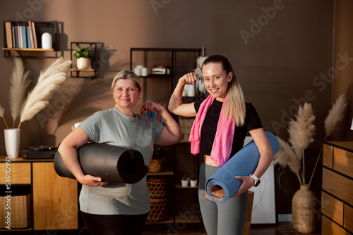 Fit women indoors showing biceps holding mats in hands looking at camera smiling. Going outside for working out together