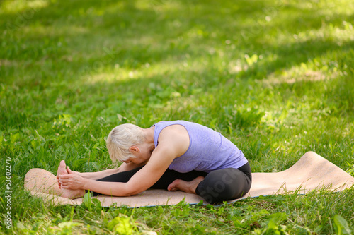 Middle-aged woman doing yoga in the park, sitting in the Janu Sirshasana exercise, Head to Knee Pose