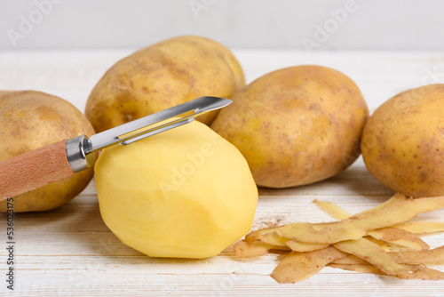 Raw peeled potato with vegetable peeler on white wooden table photo