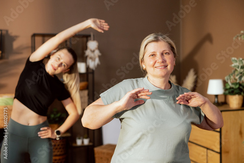 Happy elderly mother spends time with young daughter doing sport exercises stretchingindoors in modern light room looking at computer screen repeating exercises.