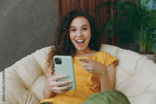 Young woman of African American ethnicity in casual clothes use point finger on mobile cell phone sits in armchair stay at home flat rest relax spend free spare time in living room indoors grey wall
