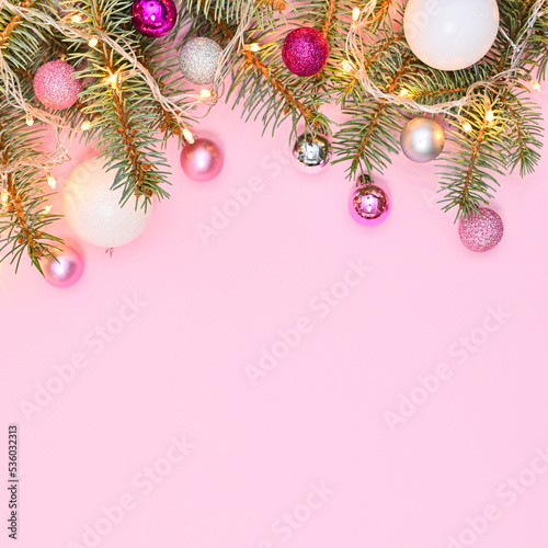 Pastel pink background with copy space and silver pine branches decorated with pink and white ornaments and lights. Flat lay