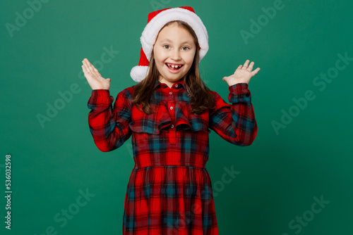 Merry surprised happy fun amazed little child kid girl 6-7 years old wear red dress Christmas hat posing spread hands look camera isolated on plain dark green background. Happy New Year 2023 concept.