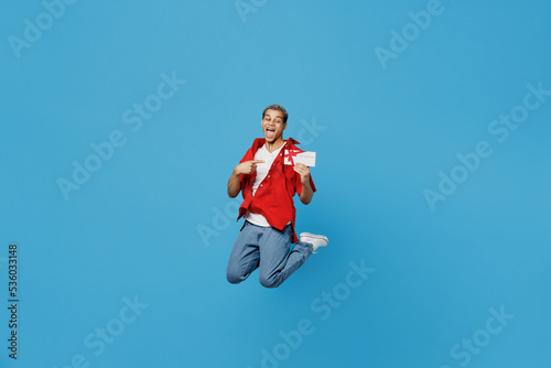 Full body satisfied happy young man of African American ethnicity in red shirt hold point finger on gift certificate coupon voucher card for store isolated on plain pastel light blue cyan background