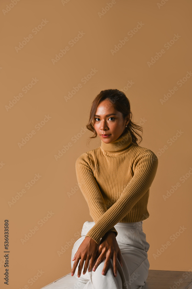 Obraz premium Fashion studio photo, Autumn and Winter concept. Attractive young woman in trendy sweater sitting over brown studio background