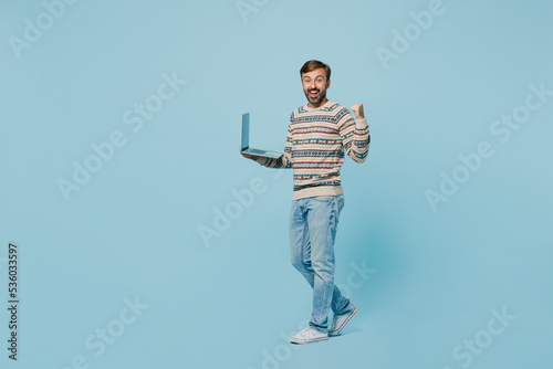 Full body side view young IT man 30s wear sweater hold use work on laptop pc computer do winner gesture isolated on plain pastel light blue cyan background studio portrait. People lifestyle concept