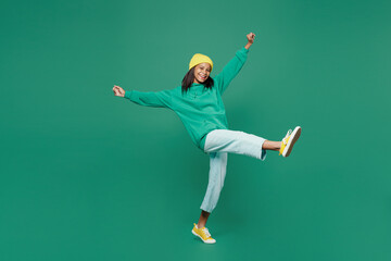 Full body side view happy little kid teen girl of African American ethnicity 13-14 years old wear casual hoody hat look camera raise up leg isolated on plain dark green background Childhood concept.