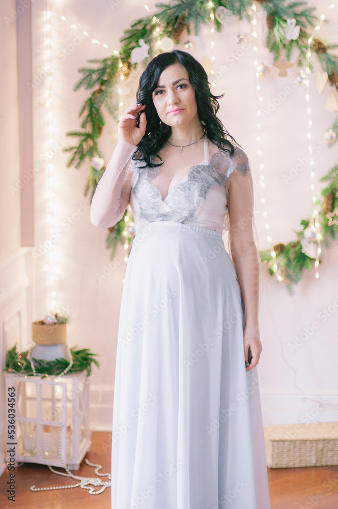 Young pregnant woman with dark hair in airy dress in a room decorated with pine needles and sparkling garlands for Christmas. Christmas mood . Pregnancy Young pregnant woman with dark hair in airy dress in a room decorated with pine needles and sparkling garlands for Christmas. Christmas mood . Pregnancy