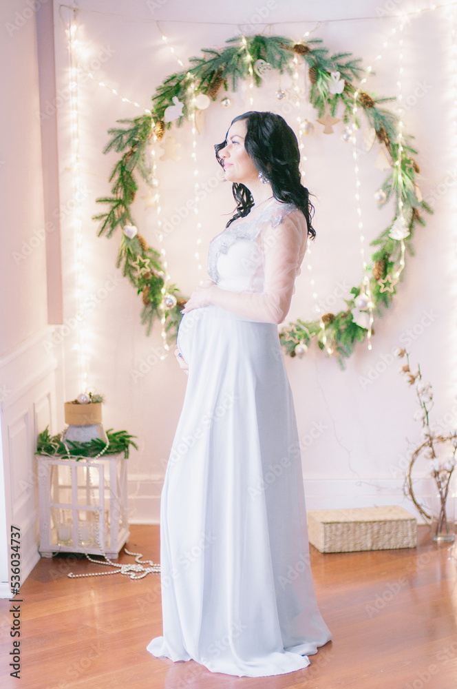 Young pregnant woman with dark hair in airy dress in a room decorated with pine needles and sparkling garlands for Christmas. Christmas mood . Pregnancy Young pregnant woman with dark hair in airy dress in a room decorated with pine needles and sparkling garlands for Christmas. Christmas mood . Pregnancy