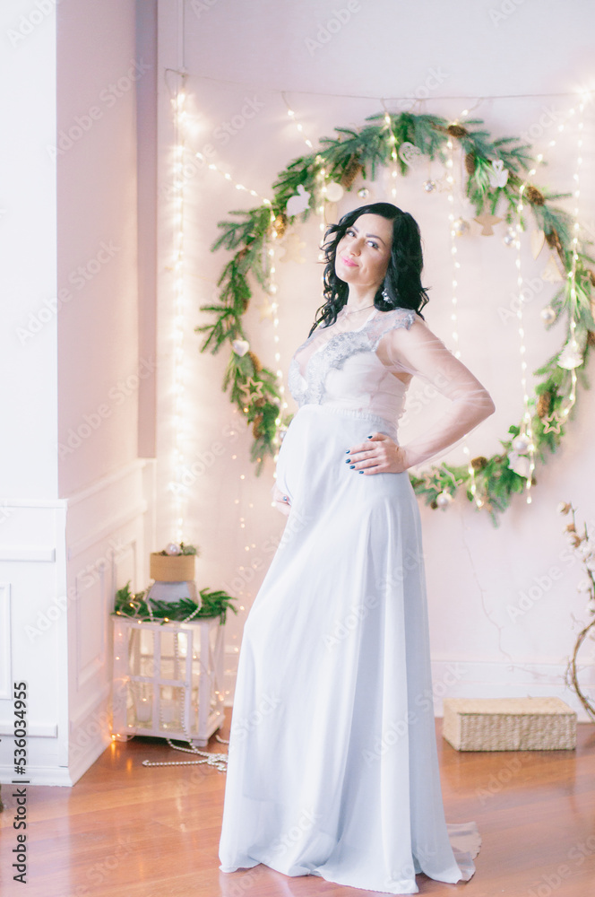 Young pregnant woman with dark hair in airy dress in a room decorated with pine needles and sparkling garlands for Christmas. Christmas mood . Pregnancy Young pregnant woman with dark hair in airy dress in a room decorated with pine needles and sparkling garlands for Christmas. Christmas mood . Pregnancy