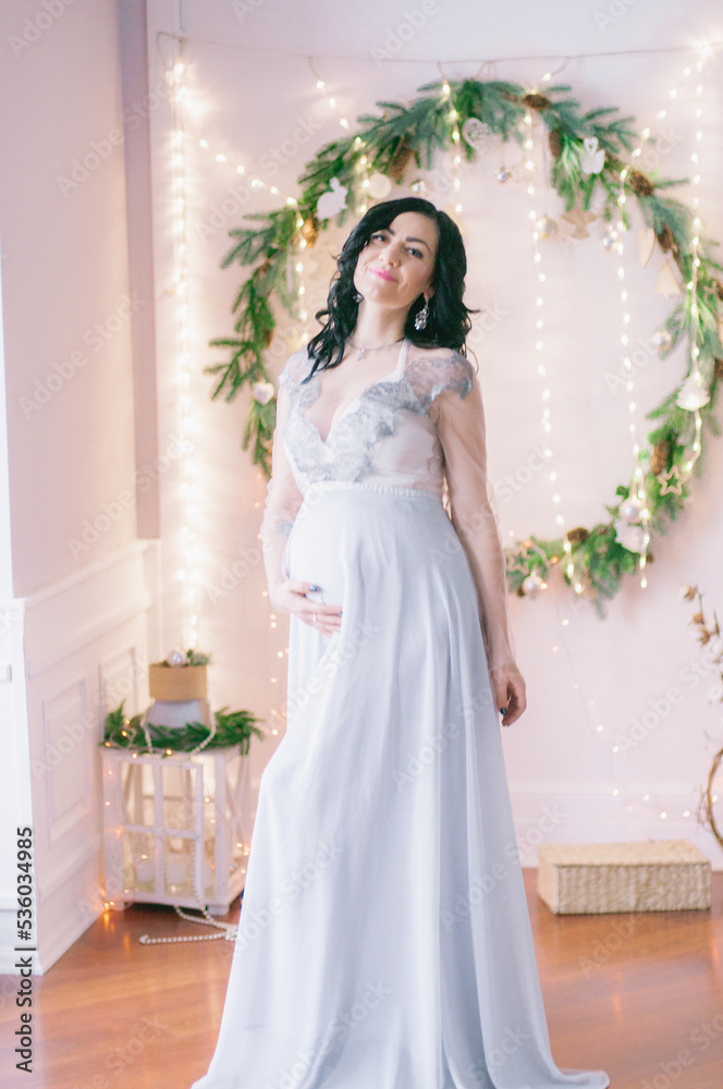 Fototapeta premium Young pregnant woman with dark hair in airy dress in a room decorated with pine needles and sparkling garlands for Christmas. Christmas mood . Pregnancy