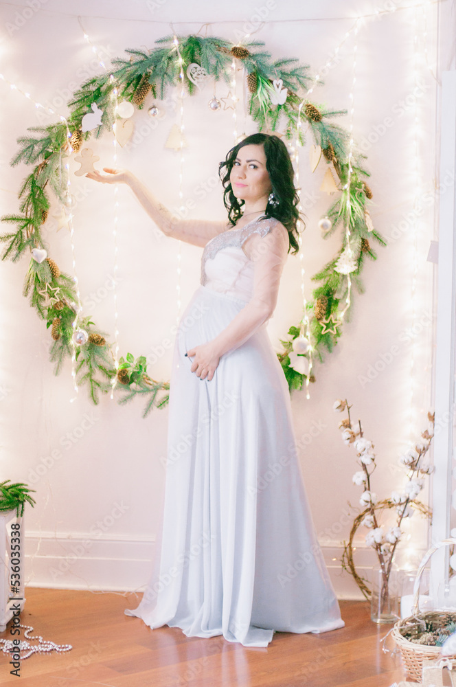Young pregnant woman with dark hair in airy dress in a room decorated with pine needles and sparkling garlands for Christmas. Christmas mood . Pregnancy Young pregnant woman with dark hair in airy dress in a room decorated with pine needles and sparkling garlands for Christmas. Christmas mood . Pregnancy