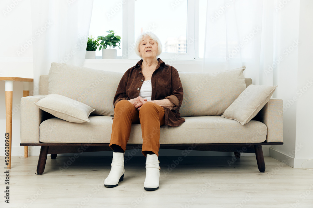 a happy elderly retired woman is sitting on a beige sofa looking at the camera with a slight smile stylishly dressed in a dark shirt and brown pants, enjoying a rest at home