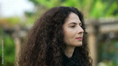 One woman standing outdoors in contemplation. Pensive thoughtful female 30s person with curly hair