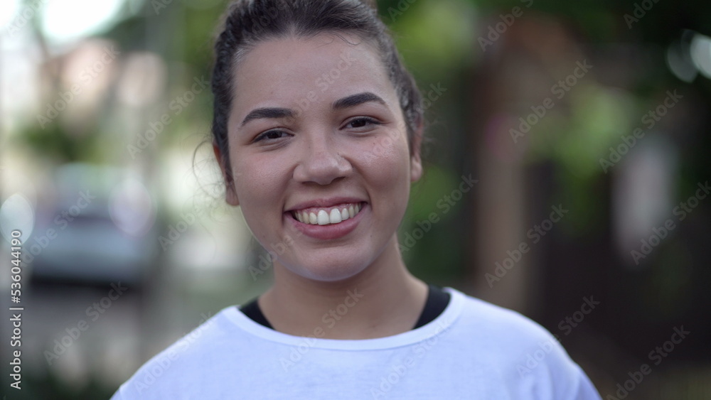 Obraz premium One Brazilian hispanic young woman walking forward toward camera. South American girl in 20s smiling in city street sidewalk