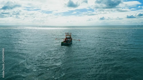 Fishing trawler, boat, ship sailing into the midday sun, trawling for fish off the Lincolnshire coast, near Skegness in the UK. showing boat pulling nets across a blue sea. Aerial, video footage photo