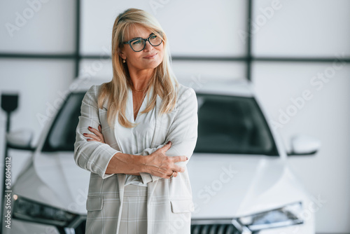 Standing with arms crossed. Woman in white formal clothes is in the car dealership