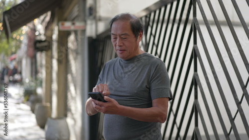 An Asian senior man using cellphone standing in city street. Older person holding smartphone device browsing internet online in daylight