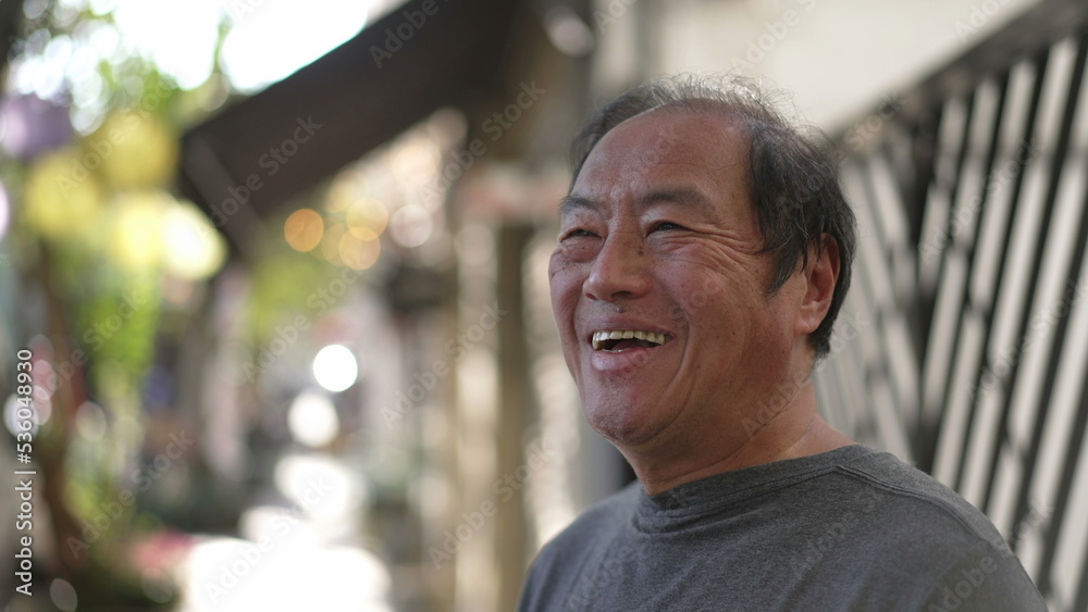 Fototapeta premium A happy Senior Asian man standing in street sidewalk smiling. Portrait of a middle aged older joyful person in daylight. Authentic real life smile