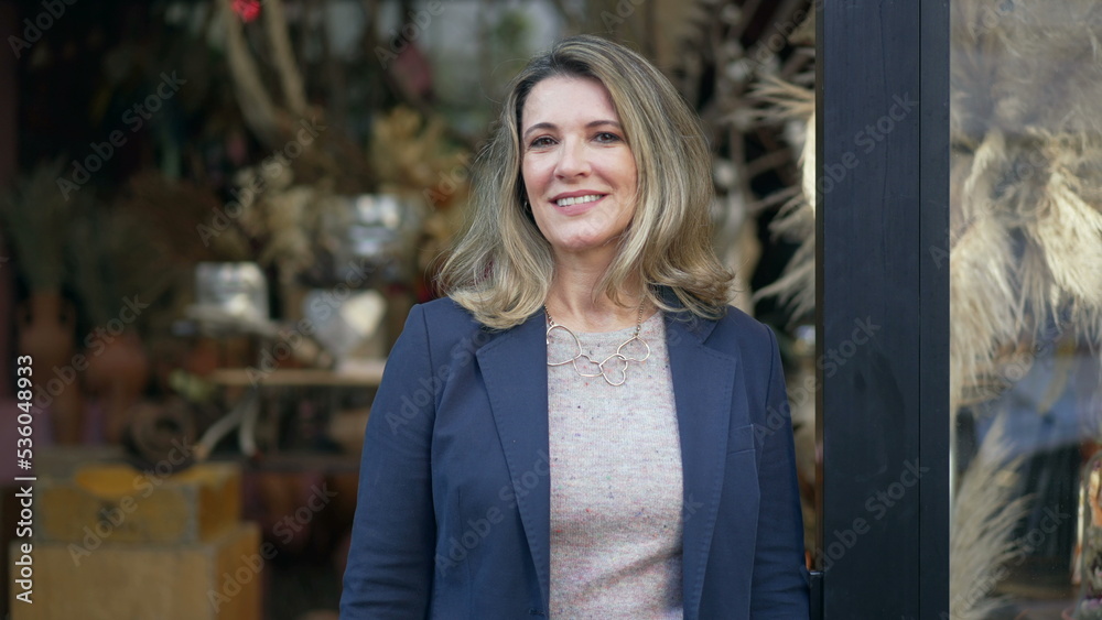 Fototapeta premium Portrait of a middle age female entrepreneur standing in front of small local business shop looking at camera