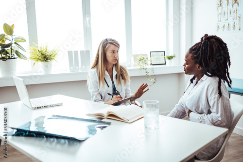 Doctor listens as a female patient discusses her health. A young female patient sits casually with her doctor as they discuss her mental health. She is seated in a chair in front of her doctor