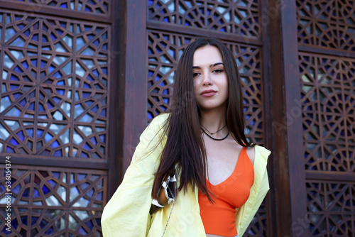 Portrait of pretty sensual girl with long hair and perfect makeup standing on background of a building with oriental patterns. Young model with handbag under the arm looking into camera, female beauty