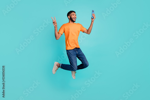 Full length photo of excited funky man dressed orange t-shirt recording video vlog modern device jumping high isolated turquoise color background