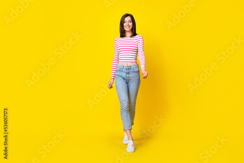 Full size photo of cute pretty cheerful woman brunette hairdo wear striped shirt jeans walking going isolated on yellow color background