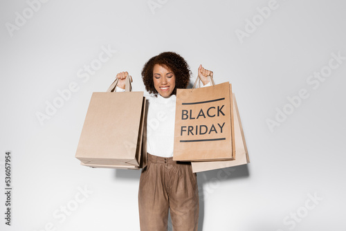 delighted african american woman with closed eyes and curly hair holding shopping bags with black friday lettering on grey. © LIGHTFIELD STUDIOS