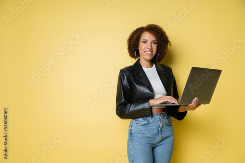 cheerful and young african american woman in leather jacket using laptop on yellow.