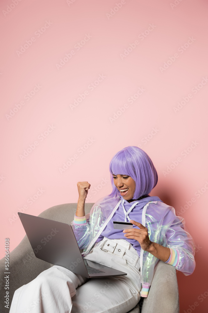 excited african american woman in purple wig using laptop while holding credit card and doing online shopping on black friday on pink. excited african american woman in purple wig using laptop while holding credit card and doing online shopping on black friday on pink.