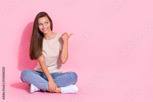 Full length photo of sweet dreamy girl dressed beige t-shirt pointing thumb empty space sitting legs crossed isolated pink color background