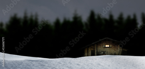 Alte Blockhütte mit beleuchtetem Fenster vor einem dunklen Tannewald im Winter